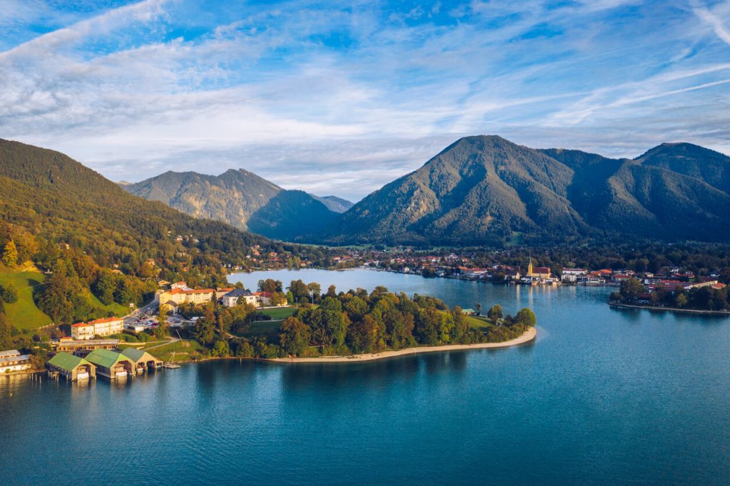 A wideshot of Lake Tegernsee, Germany, a serene lakeside town is nestled among green hills and mountains under a blue sky with wispy clouds.