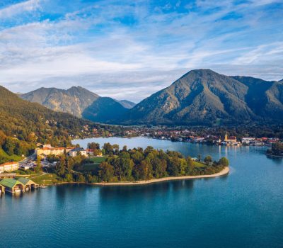 Image for A wideshot of Lake Tegernsee, Germany, a serene lakeside town is nestled among green hills and mountains under a blue sky with wispy clouds.