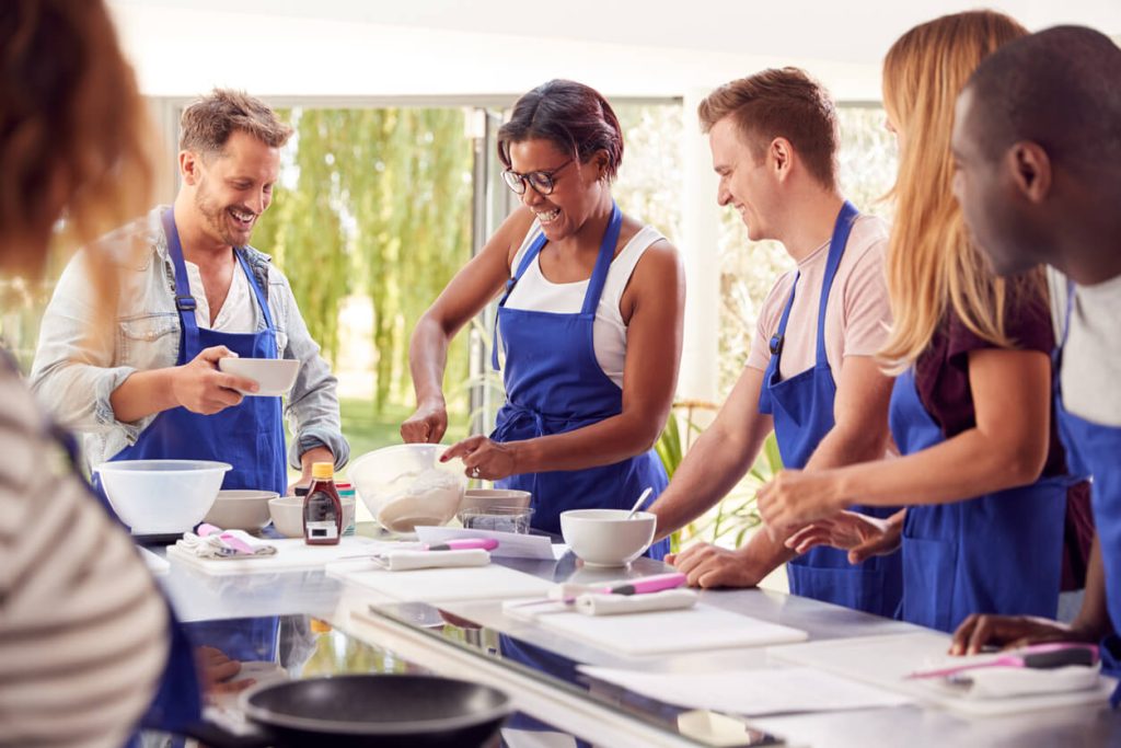 5 people in blue aprons standing around a counter during a cooking class